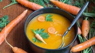 Ein schwarzer Topf mit Karottensuppe steht auf einem Holztisch. Um den Topf herum liegen Karotten.