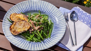 Grüner Bohnensalat und geröstetes Brot mit Bergkäse überbacken auf einem blaugemusterten Teller angerichtet und mit einer Serviette und Besteck daneben dekoriert.