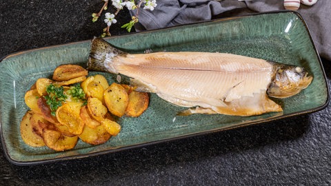 Eine selbstgeräucherte Forelle liegt schön angerichtet mit Bratkartoffeln auf einem Parzellanteller. 