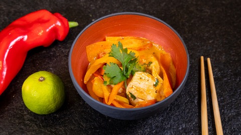 Rezept für Thai Curry: Ein leckeres Gemüsecurry mit Hähnchen und Kokosmilch in einer Schale. Rechts davon liegen Stäbchen und links davon liegen eine Limette und eine rote Paprika.