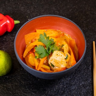 Rezept für Thai Curry: Ein leckeres Gemüsecurry mit Hähnchen und Kokosmilch in einer Schale. Rechts davon liegen Stäbchen und links davon liegen eine Limette und eine rote Paprika.