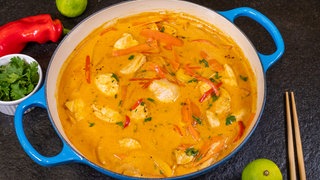 Das fertige Gericht in der Pfanne. Daneben lieben Stäbchen, Koriander in einer kleinen Schüssel, eine Limette und eine Paprika.