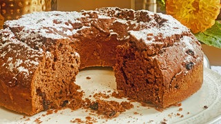Rezept für Schokokuchen mit Mandeln und Rosinen: Ein angeschnittener Schokokuchen auf einer Tortenplatte, im Hintergrund orange-farbene Blüten.
