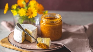 Ein Teller mit einem angeschnittenen Camembert-Käse, einem geöffneten Glas Chutney aus Früchten wie Vogelbeeren, einem Löffel mit Chutney und einem Stück Käse