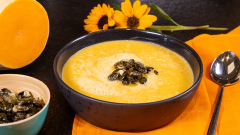 Rezept für herbstliche Kürbissuppe mit karamellisierten Kürbiskernen als Topping. Neben der Schüssel liegen ein Löffel, zwei Sonnenblumen, ein aufgeschnittener Butternus-Kürbis und eine Schale mit den Karamellisierten Kürbiskernen.