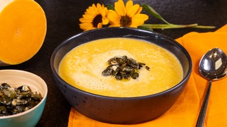 Rezept für herbstliche Kürbissuppe mit karamellisierten Kürbiskernen als Topping. Neben der Schüssel liegen ein Löffel, zwei Sonnenblumen, ein aufgeschnittener Butternus-Kürbis und eine Schale mit den Karamellisierten Kürbiskernen.