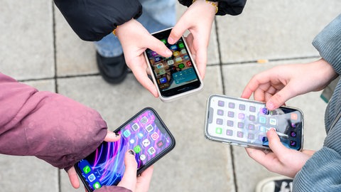 Drei Schüler mit ihren Smartphones auf dem Schulhof. Ist ein Smartphone-Verbot an Schulen wissenschaftlich betrachtet sinnvoll?