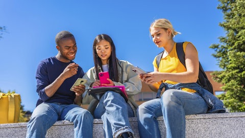 Foto einer Gruppe von Schülerinnen und Schülern die vor der Schule sitzen und Smartphones benutzen. Ein Smartphone-Verbot an Schulen ist wissenschaftlich sinnvoll.