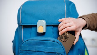 Ein Kind steckt sein Smartphone in den Schulranzen. Was sagt die Wissenschaft zum Smartphone-Verbot an Schulen.