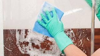Eine Frau putzt die Scheibe einer Duschkabine - Kalk entfernen