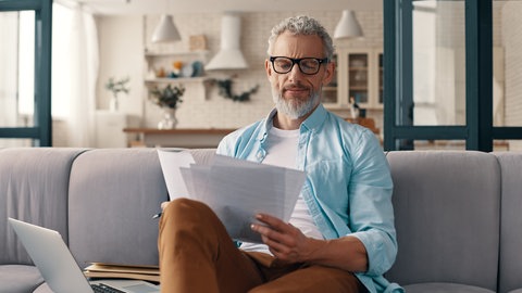 Mann sitzt mit Formularen und Laptop auf der Couch