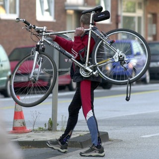 Mann trägt Fahrrad über die Straße. Immer mehr Menschen sind mit dem Rad unterwegs. Was auf dem Fahrradweg oder an Ampeln erlaubt ist. Wann es für Radfahrer Punkte in Flensburg gibt. Was beim Zubehör wichtig ist.