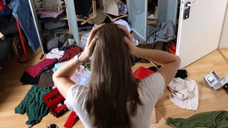 Einbruch – was tun? Eine junge Frau von hinten gesehen steht nach einem Einbruch in einer verwüsteten Wohnung und greift sich mit beiden Händen an den Kopf.