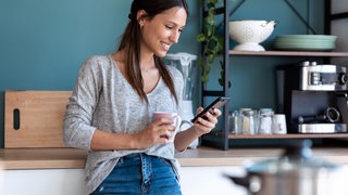 Frau steht mit dem Handy in der Küche