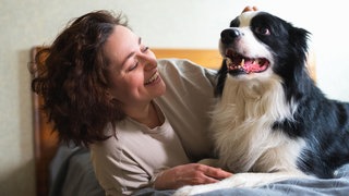 Eine Frau liegt mit ihrem Hund im Bett und lächelt ihn an. Eine neue Studie aus Wien hat untersucht, was im Hundehirn passiert, wenn Menschen lächeln.
