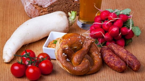 Auf einem Holztisch liegt eine typisch bayrische Brotzeit mit Radi (Rettich der Sorte "Münchner Bier"), Brot, Tomaten, einem Glas Bier, Obazda, Brezel, Radieschen und ein paar Würsten. 