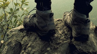 Bildausschnitt von Schuhen eines Wanderers, der auf einem schroffen Berggipfel steht, umgeben von nebliger, karger Landschaft.