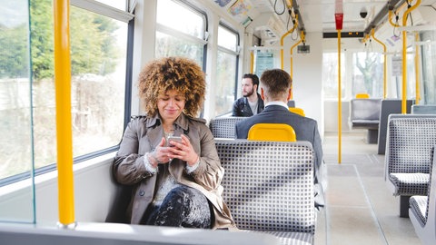 Eine Frau sitzt im Bus und schaut auf ihr Handy. Macht mit bei der ARD-Mitmachaktion #besserBahnfahren!