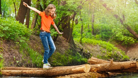 Ein Mädchen mit einem orangefarbenem T-Shirt balanciert im Wald über einen Baumstamm