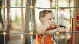 Ein traurig aussehender Junge lehnt sich auf einem Spielplatz in einer Kletternetz. 