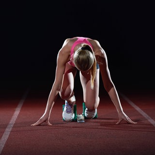 Eine Sportlerin steht am Startblock mit dunklem Hintergrund. 