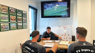 René Eberz (Videoanalyst) zeigt seinen beiden Trainer-Kollegen Julian Feit (Cheftrainer) und Martin Weber (Co-Trainer) an einer Taktiktafel, wie die Eintracht Frankfurt ihr Spiel aufzieht. René hat die Tafel neben sich. Ihm gegenüber am Tisch mit dem Rücken zur Kamera sitzen die beiden Trainer-Kollegen. Über René läuft auf einem Fernseher ein Videoausschnitt von einem Testspiel der Eintracht Frankfurt.