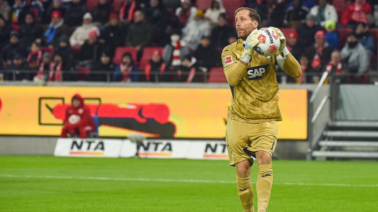 Oliver Baumann trägt ein goldenes Torwarttrikot und hält einen Fußball in der Hand