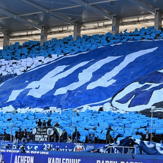 Karlsruher Fans enthüllen auf der Tribüne im Stadion ein großes KSC-Logo.