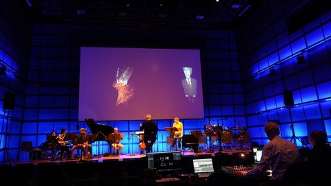 Ensemble mit Dirigent und Sängerin auf Bühne mit Leinwand im Hintergrund