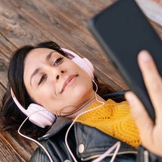 Frau mit Kopfhörern und Smartphone liegt auf dem Holzboden. Radio und Podcast hören wann und wo ihr wollt.