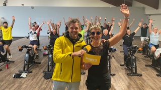 Silke und Benedikt vor dem Spinningkurs, der die Arme nach oben werfen vor Freude