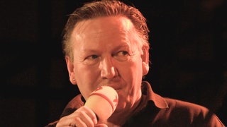 Matthias Holtmann mit Mikrofon auf der Bühne beim Finale der SWR1 Hitparade 2008.  Zum Tod von Matthias Holtmann: Wie erinnern sich Stars aus der Musikbranche, Weggefährten und SWR1 Hörer:innen an die Radiolegende und den Lebensbegleiter für viele Menschen?