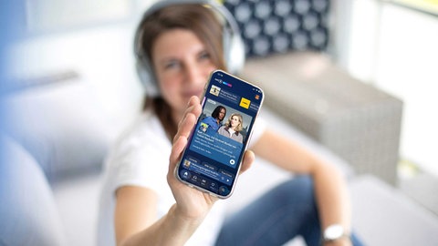 Frau im Hintergrund zeigt ein Smartphone mit der ARD Sounds App im Vordergrund.