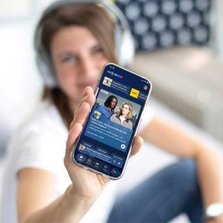 Frau im Hintergrund zeigt ein Smartphone mit der ARD Sounds App im Vordergrund.