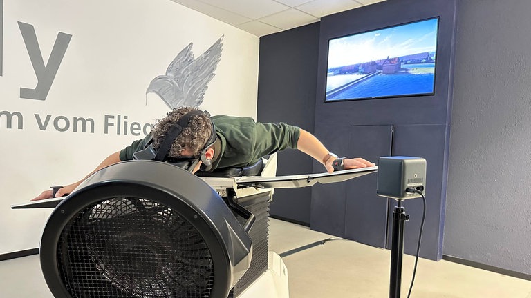 SWR1 Moderator Julian Hammerstein auf dem Vogelflugsimulator "Birdly". Auf dem Bildschirm im Hintergrund ist das virtuelle Bild des Vogelflugs zu sehen.