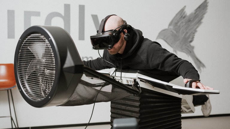 Ein Mann liegt auf dem Vogelflugsimulator "Birdly". Er trägt eine VR-Brille und Kopfhörer.