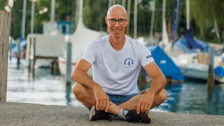Atlantik-Soloruderer Martin Stengele sitzt im Schneidersitz, im Hintergrund sind Boote sichtbar. Martin Stengele versuchte alleine quer über den Atlantik zu rudern.