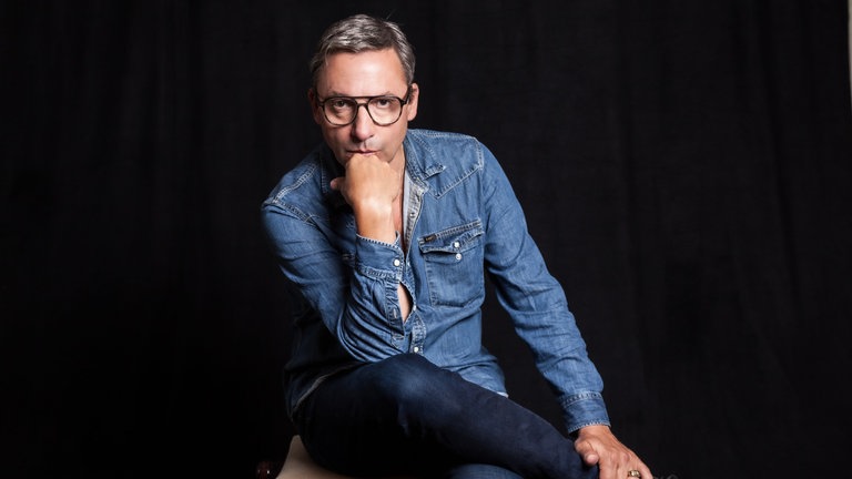 Musiker und Schriftsteller Rocko Schamoni sitzt mit übergeschlagenem Bein und auf die Hand aufgestütztem Kopf auf einem Stuhl. Die "Rocko Schamoni Supershow" trendet in der ARD Mediathek, seine "Dorfpunks" wurden zum Kultbuch, 20 Jahre später kommt mit "Pudels Kern" die Fortsetzung. Rocko Schamoni nimmt uns in SWR1 Leute mit in die 80er und 90er, zu den Toten Hosen, Bela B., Peter Maffay, Helge Schneider und in den legendären Hamburger Pudelclub.