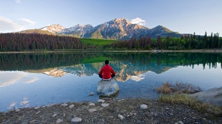 Ein Mann sitzt in einem See auf einem großen Stein und blickt auf ein Bergpanorama. In SWR1 Leute spricht Wolfgang Fasser über die große Bedeutung von Ruhe und Stille für uns.