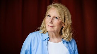 Die Schauspielerin und Kabarettistin Maren Kroymann steht vor einem roten Vorhang und schaut in die Kamera.
