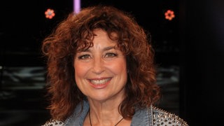 Schauspielerin Isabel Varell sitzt in einer Jeansjacke auf einem Stuhl und lacht in die Kamera.