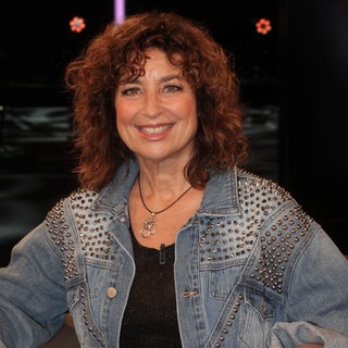 Schauspielerin Isabel Varell sitzt in einer Jeansjacke auf einem Stuhl und lacht in die Kamera.