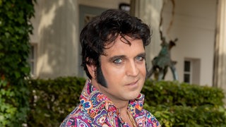 Nils Strassburg als Elvis in einem bunten Hemd und mit goldenen Ringen und schwarzen Koteletten. Er schaut direkt in die Kamera.