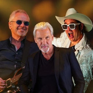 Drei Musiker stehen vor einem warm verschwommenen Hintergrund. Links der Sänger von Bitch is Back mit weißem Cowboyhut, weißer Fransenjacke und großer Sonnenbrille in einer energiegeladenen Pose, in der Mitte Johnny Logan in dunklem Sakko, rechts Nik Kershaw mit runder Sonnenbrille und E-Gitarre.