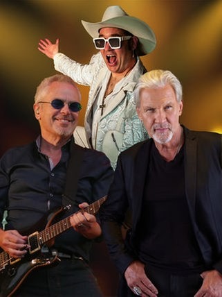 Drei Musiker stehen vor einem warm verschwommenen Hintergrund. Links der Sänger von Bitch is Back mit weißem Cowboyhut, weißer Fransenjacke und großer Sonnenbrille in einer energiegeladenen Pose, in der Mitte Johnny Logan in dunklem Sakko, rechts Nik Kershaw mit runder Sonnenbrille und E-Gitarre.