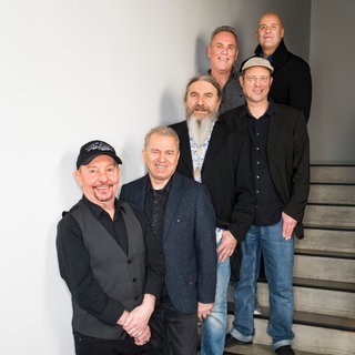 Die Band steht hintereinander auf einer Treppe und sie schauen alle in die Kamera.