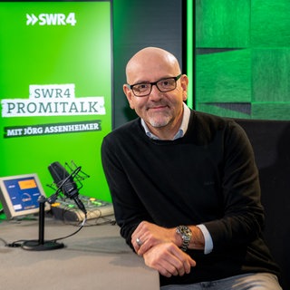 Moderator Jörg Assenheimer sitzt lächelnd im Studio am Tisch mit zwei Mikros und Bildschrim, im Hintergrund das Logo des SWR4 Promitalk auf einem Monitor.