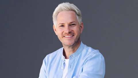 SWR4 Moderator Marcel Wagner mit offenem, blauen Hemd, darunter einem weißen T-Shirt lächelt in die Kamera.