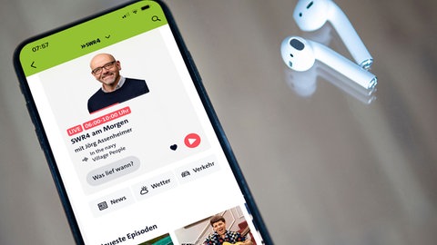 Ein Smartphone mit geöffneter SWR4 Wellenheimat in der Audiothek liegt auf dem Tisch, daneben In-Ear-Kopfhörer