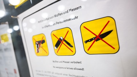 Auf einer U-Bahn-Hinweistafel wird mit drei Piktorgrammen, auf denen eine durchgestrichene Pistole, ein durchgestrichenes Taschen- und ein durchgestrichenes Küchenmesser zu sehen sind, auf das Verbot des Mitführens von Waffen und Messern hingewiesen.